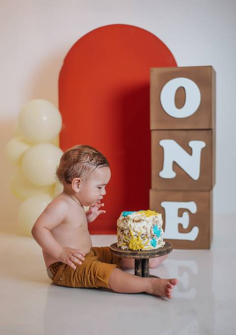 Bebê observando o bolo no ensaio Smash the Cake de 1 ano, fotografia infantil em estúdio em Farroupilha, com cenário em tons quentes.'