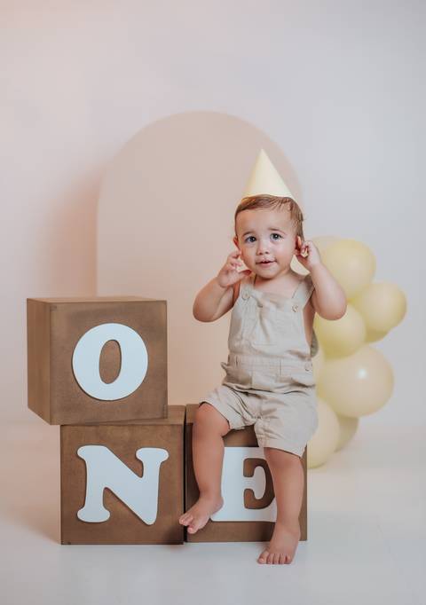Bebê de 1 ano usando chapéu de aniversário sentado ao lado de cubo decorativo em ensaio infantil'
