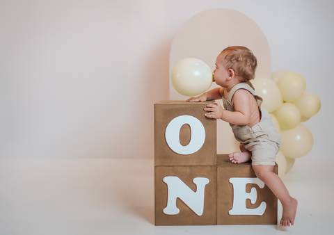 Bebê de 1 ano sentado ao lado de cubos com a palavra ONE e balões claros em estúdio infantil'