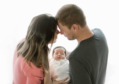 Ensaio newborn tradicional com o Ravi nos primeiros dias de vida, bebê no colo dos pais em estúdio com fundo claro'