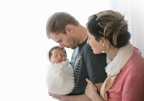 Fotografia newborn tradicional com bebê recém-nascido no colo dos pais, registro familiar em estúdio'