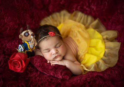 Bebê recém-nascida em ensaio newborn temático A Bela e a Fera, figurino amarelo e cenário vermelho em estúdio'