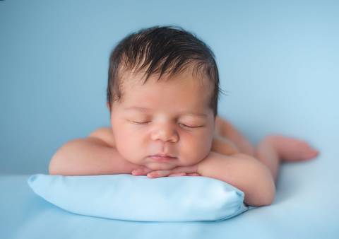 Ensaio newborn tradicional com bebê recém-nascido apoiado em almofada azul, cenário minimalista'