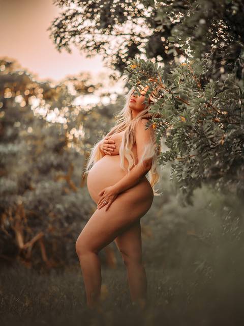 Mulher grávida em ensaio gestante nu, em meio à natureza, com a barriga em evidência e postura serena. Registro do corpo feminino durante a gestação sem conotação sexual.'