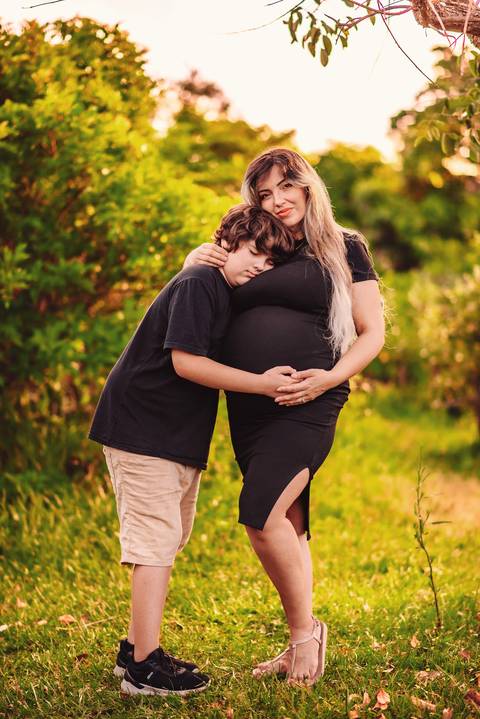 Mulher grávida vestida de preto em ensaio gestante, com o filho mais velho abraçando a barriga, em cenário natural, representando afeto e conexão familiar.'