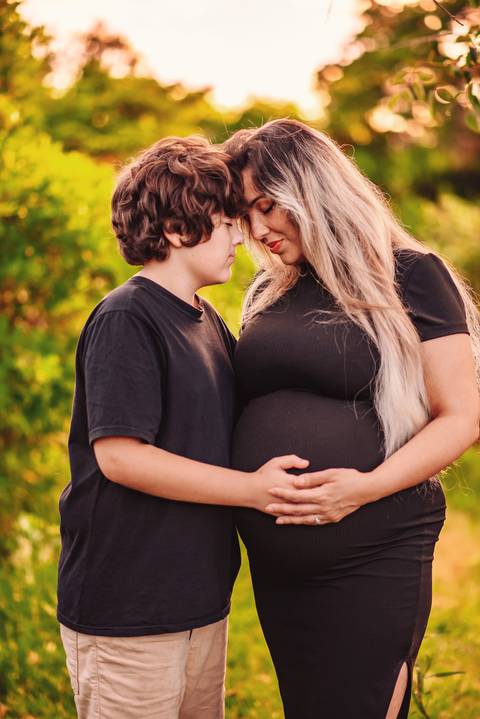 Mulher grávida e filho mais velho em ensaio gestante ao ar livre, com as mãos apoiadas sobre a barriga, simbolizando a chegada de um novo membro da família.'