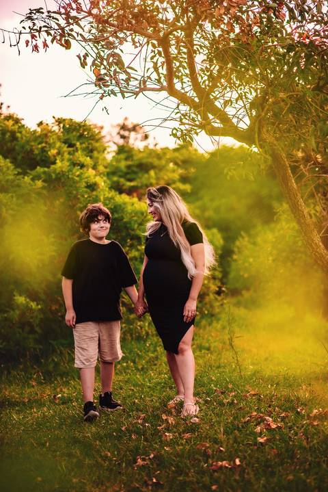 Mulher grávida e filho em ensaio gestante artístico ao ar livre, de mãos dadas, em ambiente natural, registrando a gestação dentro do contexto familiar.'