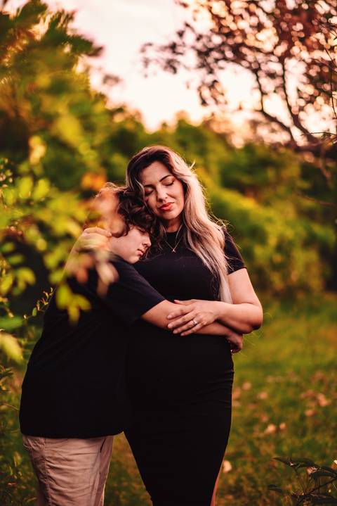 Mulher grávida em ensaio gestante, abraçada ao filho mais velho, em cenário natural, com foco na relação mãe e filho durante a gestação.'