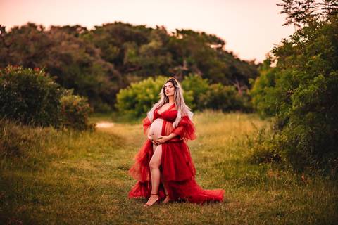 Mulher grávida em ensaio gestante artístico ao ar livre, vestindo vestido vermelho, com a barriga em evidência, em meio à natureza.'