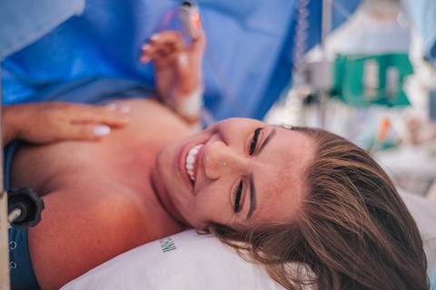 Mãe sorrindo após o nascimento do bebê, em momento de alívio e emoção no pós-parto imediato.'