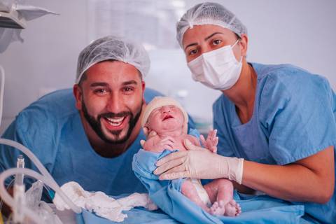 Pai sorrindo ao lado do recém-nascido logo após o nascimento, acompanhado pela equipe médica em sala de parto hospitalar.'