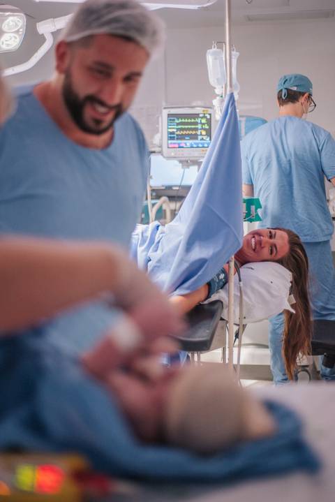 Mãe sendo conduzida pela equipe médica após o parto, ainda na sala de nascimento do hospital.'