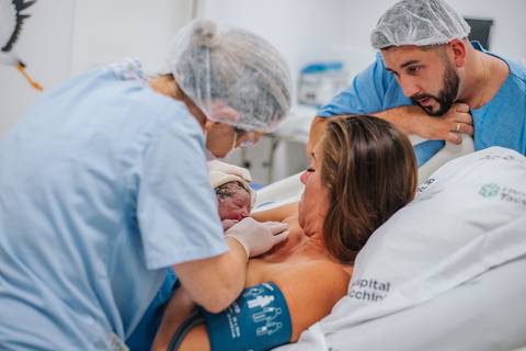 Mãe e pai observando os primeiros cuidados com o recém-nascido em sala de parto hospitalar.'