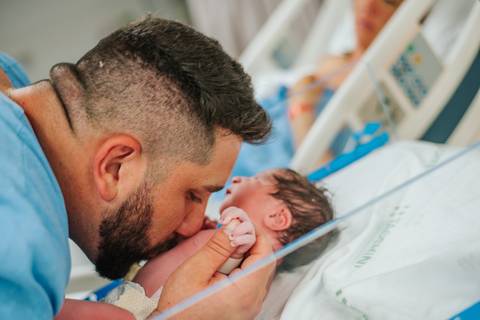 Pai beijando o recém-nascido nos primeiros momentos de vida, em ambiente hospitalar.'