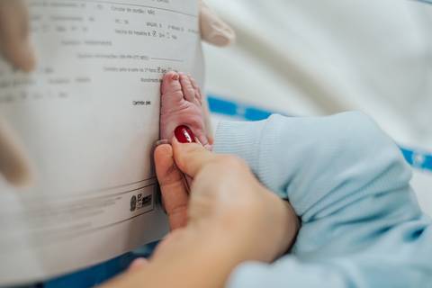 Mãe assinando documentos hospitalares enquanto segura a mão do recém-nascido após o parto.'