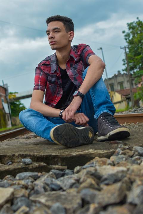 garoto sentado no trilho do trem com uma mão segurando o punho contrário, de camisa xadrez e calça jeans.'