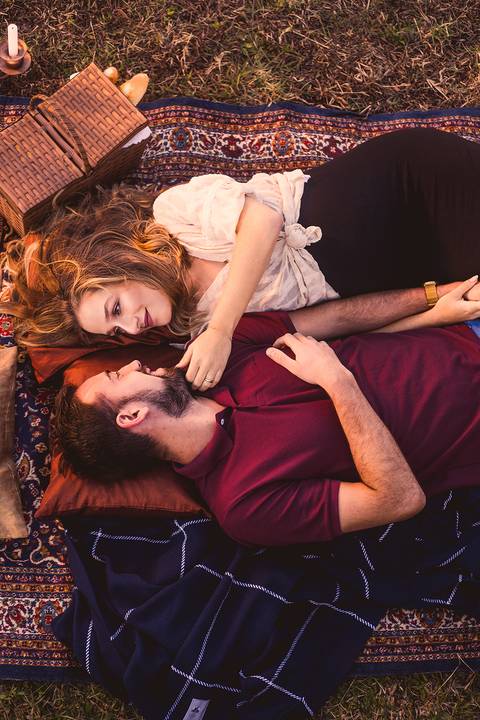 Retrato tirado de cima, de casal deitado em tapete na grama, com almofadas e cenário em tons quentes'