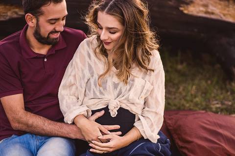 Retrato de casal gestante esperando bebe, sentados em tapete persa na grama com tronco ao fundo'