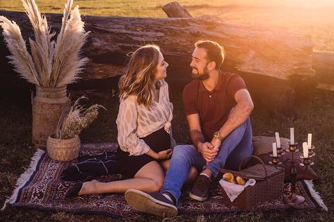 Retrato de casal sentado em cenário em tons pasteis, na grama, no por do sol, com a luz bem amarelada ao fundo'