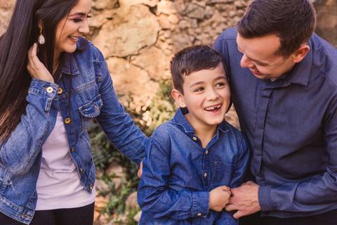 Mãe, pai e filho brincando, sorrindo, com parede de pedras ao fundo. Retrato família alegre e cheio de carinho'