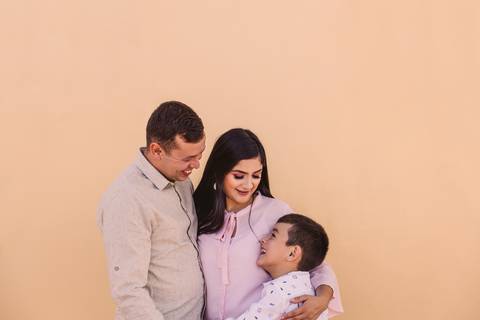 Pai, mãe e filho, abraçados em frente a parede amarela pastel. Muito carinho em família'