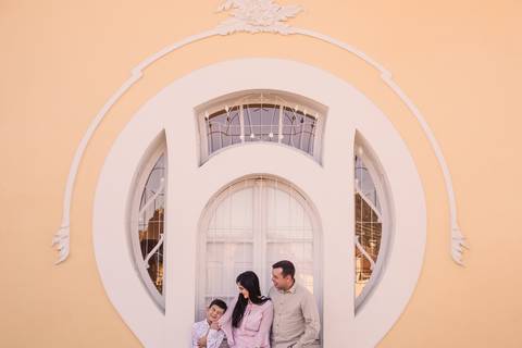 Filho, mãe e pai, brincando e sorrindo, emoldurados por uma porta branca de design antigo.'