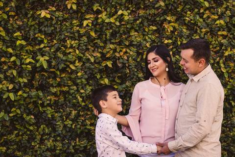 Filho, mãe e pai, em frente a parede de plantas. Sorrindo, carinhosamente.'