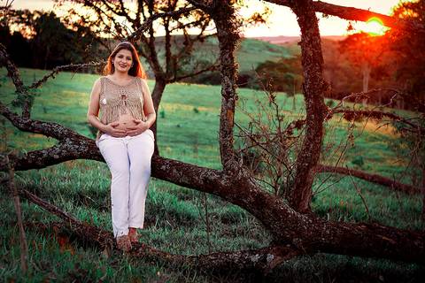 gravida sorrindo sentada em arvore em ensaio de gestante no por do sol na zona rural de mandaguari regiao de maringa'