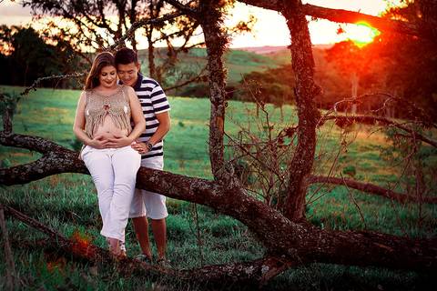 casal sorrindo sentados em arvore em ensaio de gestante no por do sol na zona rural de mandaguari na regiao de maringa'