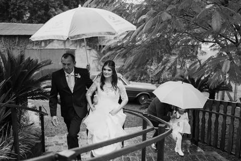 Noiva chegando com o pai sob guarda-chuva em dia de chuva, casamento em Lajeado na Casa Irmando, fotografia por Alba Estúdio.'
