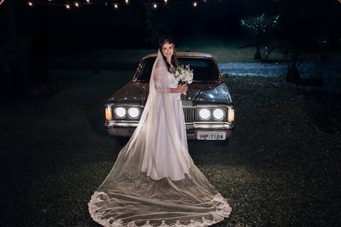 Noiva posa com buquê em frente a um carro antigo à noite, com véu esvoaçante e luzes ao fundo. Casamento na Casa Irmando em Lajeado. Foto por Alba Estúdio.'