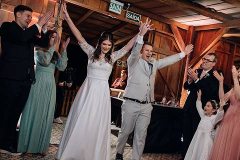 Entrada dos noivos na recepção com braços erguidos e alegria contagiante, rodeados por convidados. Casamento na Casa Irmando em Lajeado. Foto por Alba Estúdio.'