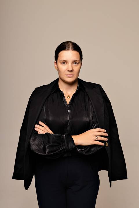 Mulher de pé com blazer escuro por cima dos ombros, camisa preta, olhar direto, fundo bege minimalista'