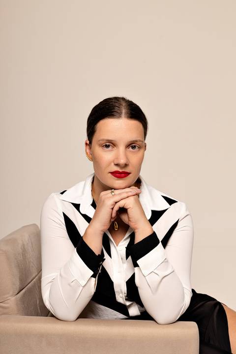 Mulher sentada de lado em sofá claro, blouse preta acetinada, olhar sereno, cenário clean'