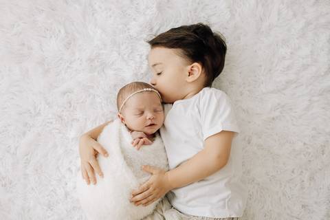 “Irmãos deitados lado a lado em tapete felpudo durante ensaio newborn”'