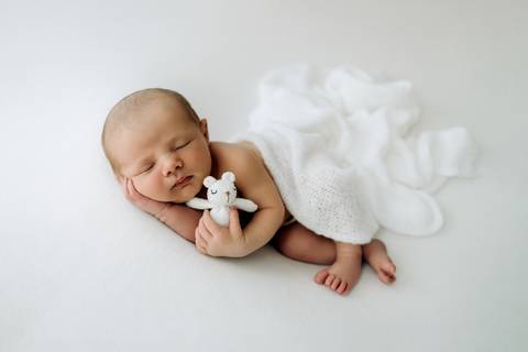 Bebê Lorenzo deitado em manta branca segurando mini ursinho de pelúcia, fotografia newborn no Alba Estúdio.'