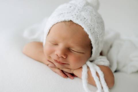 Bebê Lorenzo sorrindo levemente enquanto dorme, usando gorro branco de tricô, ensaio newborn no Alba Estúdio.'
