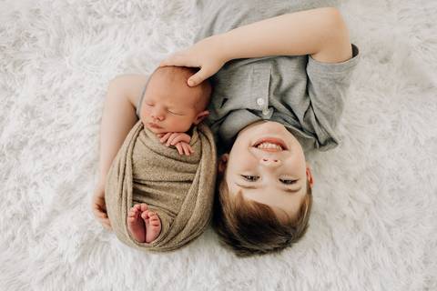 Menino deitado ao lado do irmão recém-nascido Lorenzo enrolado em manta bege, em ensaio newborn no Alba Estúdio.'