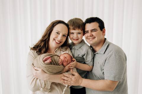Retrato em família no ensaio newborn de Lorenzo, bebê enrolado em manta bege no colo da mãe, junto ao pai e irmão mais velho, fotografia no Alba Estúdio.'