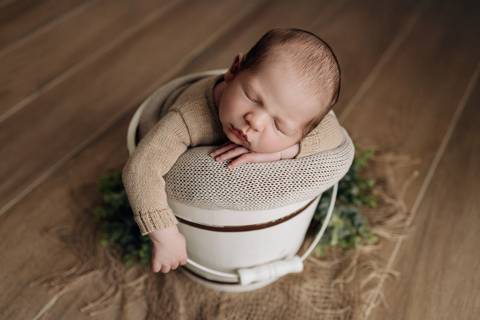 Foto newborn em Lajeado – bebê dormindo dentro de balde branco, vestido com roupa de tricô bege, fotografia feita no Alba Estúdio.'