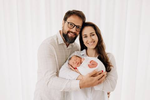 Ensaio newborn em família em Lajeado – mãe e pai segurando recém-nascida enrolada em manta branca, fotografia feita no Alba Estúdio.'