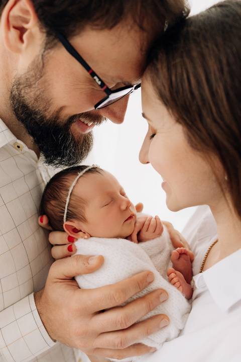 Pais sorridentes seguram a bebê Maria Helena, enrolada em manta branca, durante ensaio newborn no Alba Estúdio em Lajeado.'