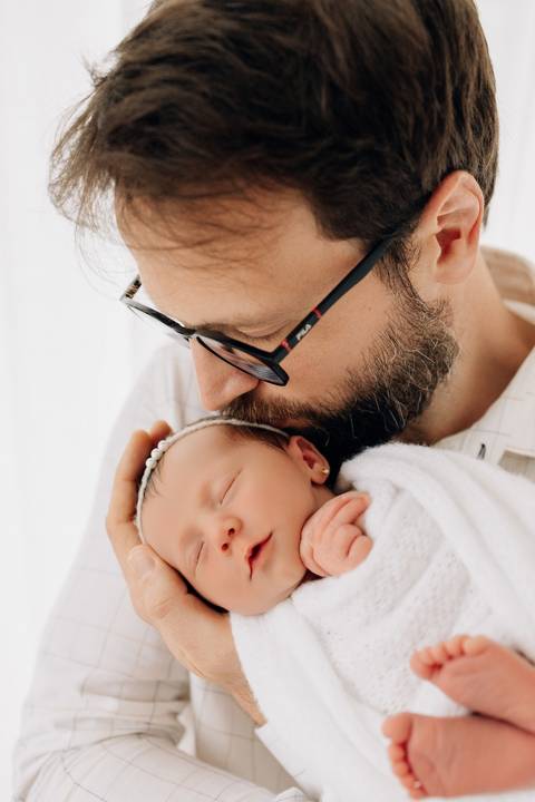 Pai beija com carinho a filha recém-nascida, enrolada em manta branca, durante ensaio newborn no Alba Estúdio em Lajeado.'