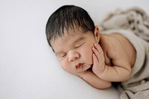 Ensaio newborn do bebê Noah dormindo de bruços com a mão apoiada no rosto, coberto por manta bege no estúdio Alba Estúdio em Lajeado.'