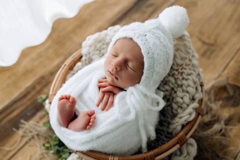 Bebê Oliver dormindo enrolado em manta branca dentro de cesto, usando gorro de tricô branco com pompom, no estúdio Alba Estúdio em Lajeado.'