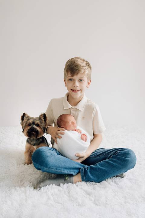 Bebê Oliver no ensaio newborn, no colo do irmão mais velho com um cachorro ao lado, no estúdio Alba Estúdio em Lajeado.'