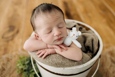 Fotografia newborn em Lajeado – bebê recém-nascido dormindo dentro de balde branco, abraçado a ursinho de crochê branco. Registro realizado pelo Alba Estúdio.'