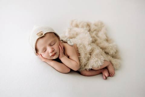 Ensaio newborn em Lajeado – bebê deitado de lado sobre fundo branco, usando gorro bege com botão, coberto por manta felpuda, em pose clássica de estúdio. Foto do Alba Estúdio.'