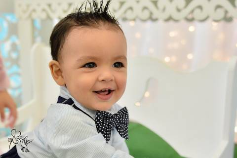 Eduardo sorri com calça azul,camisa branca e gravata borboleta azul no seu aniversário de um ano, by Eli Matos registrou'