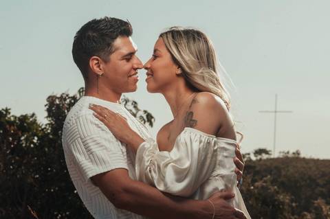 dunas de areia - ensaio de casal   - casal quase se beijando  no por so sol - noivo camiseta bege e noiva com blusa decotada  com uma cruz no fundo '